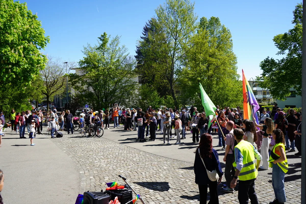 Man sieht Menschen auf einem Platz versammelt. Ebenfalls sichtbar ist eine grosse Regenbogenfahne.
